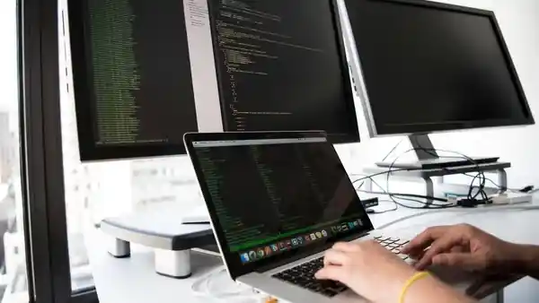 Hands typing code on a laptop beside two monitors.